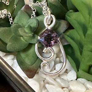 May include: A silver pendant necklace with a purple gemstone set in a square shape. The pendant is attached to a silver chain and has a delicate, ornate design.