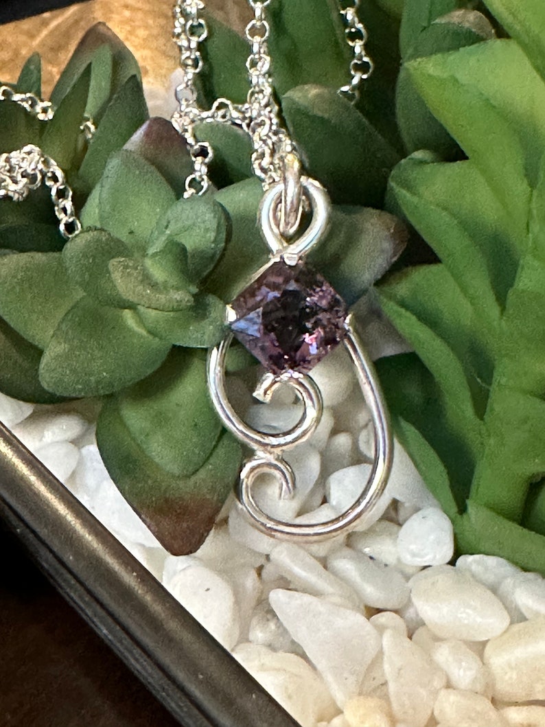 May include: A silver pendant necklace featuring a purple gemstone set in a square shape. The pendant is attached to a silver chain and has a delicate, ornate design.
