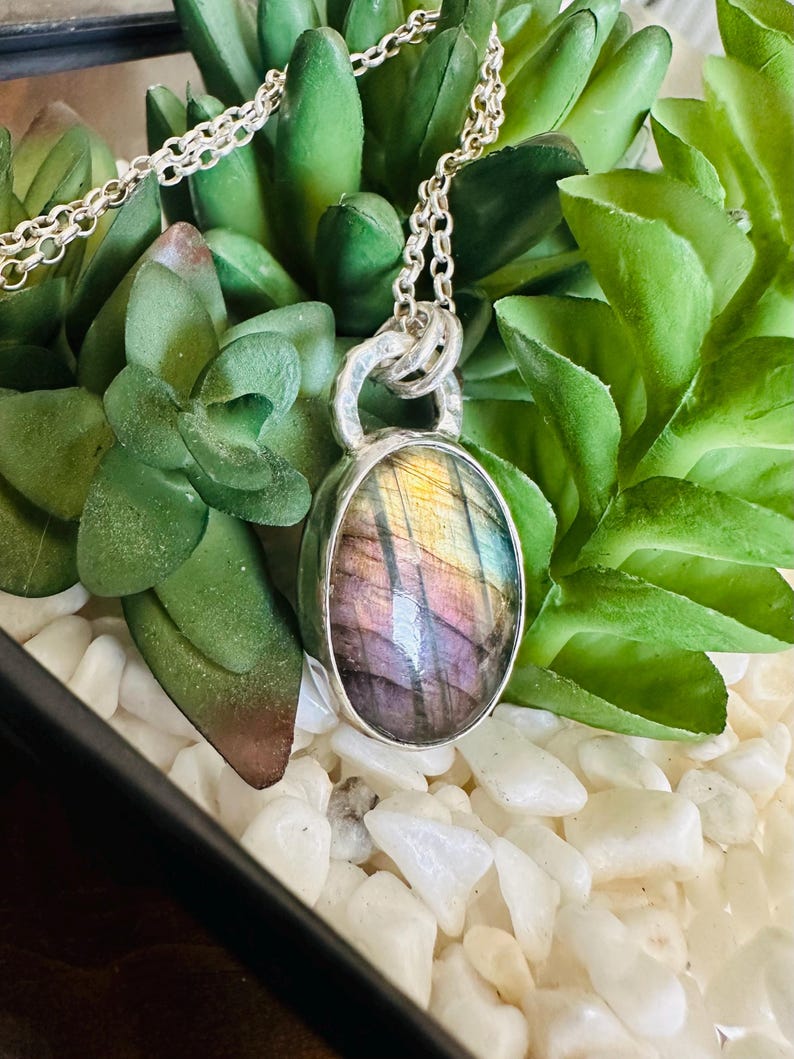 May include: A silver necklace with an oval pendant containing an iridescent gemstone. The stone shows purple, yellow, and blue colours. The necklace is set against green succulents and white pebbles.
