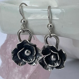 May include: A pair of silver flower-shaped earrings with a textured finish. The earrings are hanging from a silver hook.