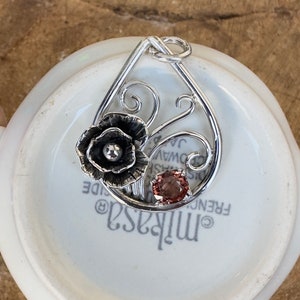 May include: A silver pendant featuring a delicate floral design with a pink gemstone. The pendant is shaped like a teardrop and has a swirl pattern.
