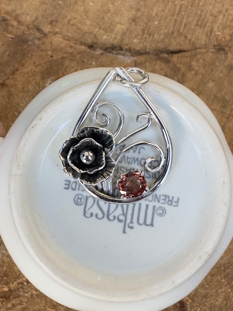 May include: A silver pendant featuring a delicate floral design with a pink gemstone. The pendant is shaped like a teardrop and has a swirl pattern.