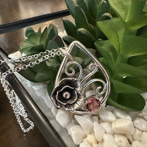 May include: A silver pendant necklace with a delicate floral design and a pink gemstone. The pendant is shaped like a teardrop and features a stylized flower with a pearl-like center. The necklace is on a silver chain.