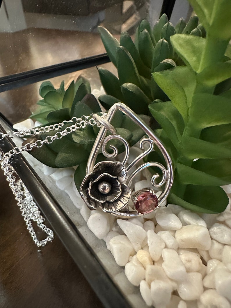 May include: A silver pendant necklace with a delicate floral design and a pink gemstone. The pendant is shaped like a teardrop and features a stylized flower with a pearl-like center. The necklace is on a silver chain.