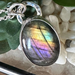 May include: A silver pendant necklace with an oval labradorite stone. The stone exhibits a vibrant play of colour, including yellow, green, purple, and blue. The pendant hangs from a delicate silver chain, showcasing a unique and eye-catching design.