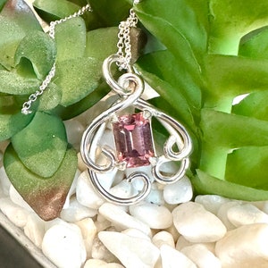 May include: A silver pendant necklace with a pink gemstone set in a delicate, swirling design.