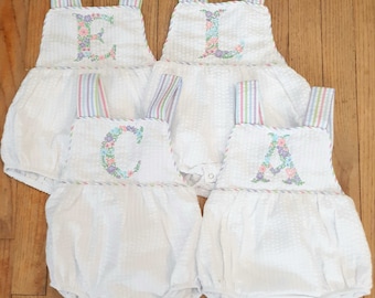 May include: Four white baby onesies with colorful striped straps. Each onesie has a different letter embroidered on the front with floral accents. The letters spell out "ELCA".