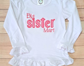 May include: White long-sleeved shirt with a pink and white gingham "Big Sister" design and the name "Mari" embroidered on the front. The shirt has ruffles on the sleeves and bottom.