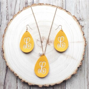 May include: A set of yellow leather earrings and a necklace with a pearl accent. Each piece features a white embroidered letter "L" in a cursive font.