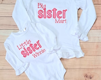 May include: Two white long-sleeved shirts with red and white checkered lettering. The larger shirt says "Big Sister Mari" and the smaller shirt says "Little Sister Winnie".