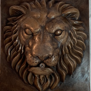 Può includere: Una scultura in bronzo di una testa di leone con una criniera dettagliata e una superficie testurizzata. La bocca del leone è leggermente aperta, rivelando un piccolo foro al centro della sua lingua.
