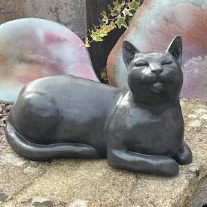 May include: A dark grey cat sculpture in a relaxed pose, with closed eyes and a slight smile. The statue is on a textured stone surface, with blurred, colourful decorative stones in the background. The cat's fur appears smooth and glossy.