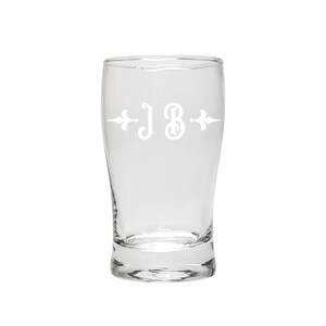 Op de afbeelding: Een helder glazen bierglas met een wit gegraveerd ontwerp van de initialen "JB" met decoratieve versieringen.