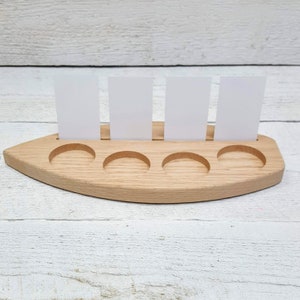 May include: Wooden boat-shaped card holder with five slots for cards. The holder is made of light brown wood and has a natural finish. There are four white cards standing upright in the slots.