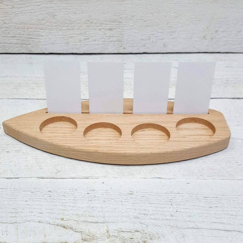 May include: Wooden boat-shaped card holder with five slots for cards. The holder is made of light brown wood and has a natural finish. There are four white cards standing upright in the slots.