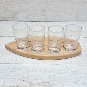 May include: A wooden serving tray with four clear glass shot glasses. The tray is shaped like a boat and has a natural wood finish.