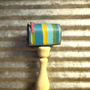 May include: A miniature mailbox with blue, yellow, and green stripes and a pink roof sits on top of a wooden pedestal. The mailbox and pedestal are in front of a corrugated metal background.