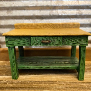 May include: A small green wooden table with a single drawer and a shelf. The table top is a light brown wood.