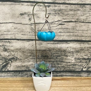 May include: A decorative plant stand featuring a small, turquoise hanging planter suspended above a white square pot holding a succulent. The stand is made of a dark metal and is set against a wooden background.