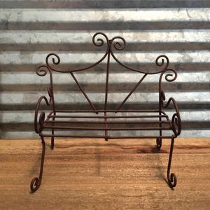 May include: A small, rustic, metal garden bench with a scrolled design. The bench is made of wire and has a slightly rusted finish.
