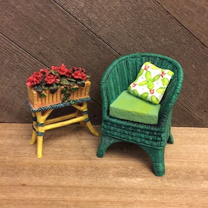 May include: Miniature wicker chair and plant stand set. The green chair has a green cushion and a cactus-print pillow. The yellow plant stand holds a basket of red flowers.