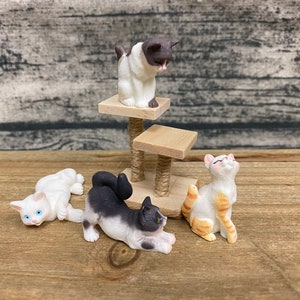 May include: A collection of miniature cat figurines in various poses. One cat is on a small cat tree, while others are lying down or sitting. The cats are white, black, and orange, with detailed features.