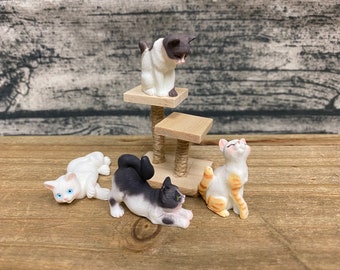 May include: Miniature cat figurines in different poses, including one on a small cat tree. The cats are white, black, and orange, with detailed features. The figurines are set on a wooden surface.