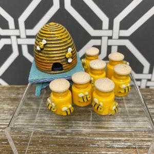 May include: A set of eight small honey jars with bee designs, including a ceramic beehive with a yellow and brown striped pattern.
