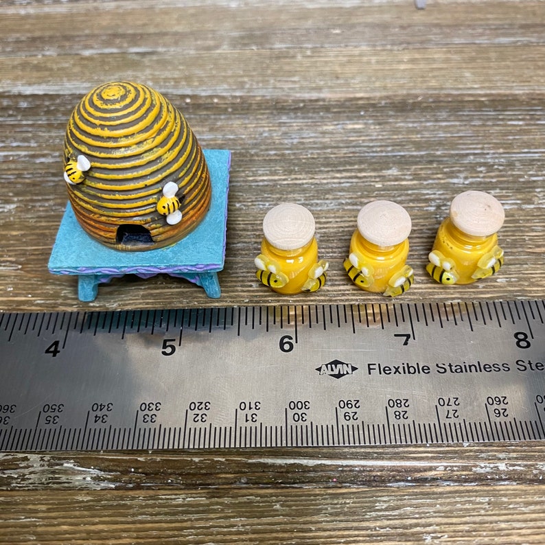 May include: A miniature beehive with a wooden base and three small jars of honey, each with a bee on top. The jars are yellow with a brown lid and the bees are black and yellow. The beehive is brown and yellow with a small opening in the front. The scene is set on a wooden surface with a ruler for scale.