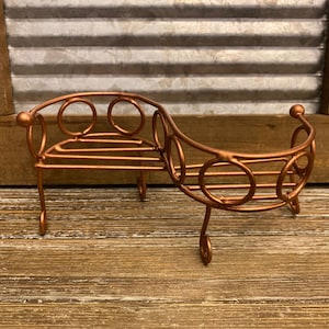 May include: A small, decorative copper-colored metal bench with a curved design and circular accents. The bench is made of wire and has a rustic look.