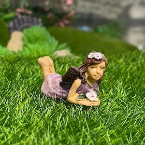 May include: A miniature fairy figurine with brown hair and a flower crown, wearing a purple dress and holding a bouquet of white flowers. The fairy is lying on its side on a bed of green grass.