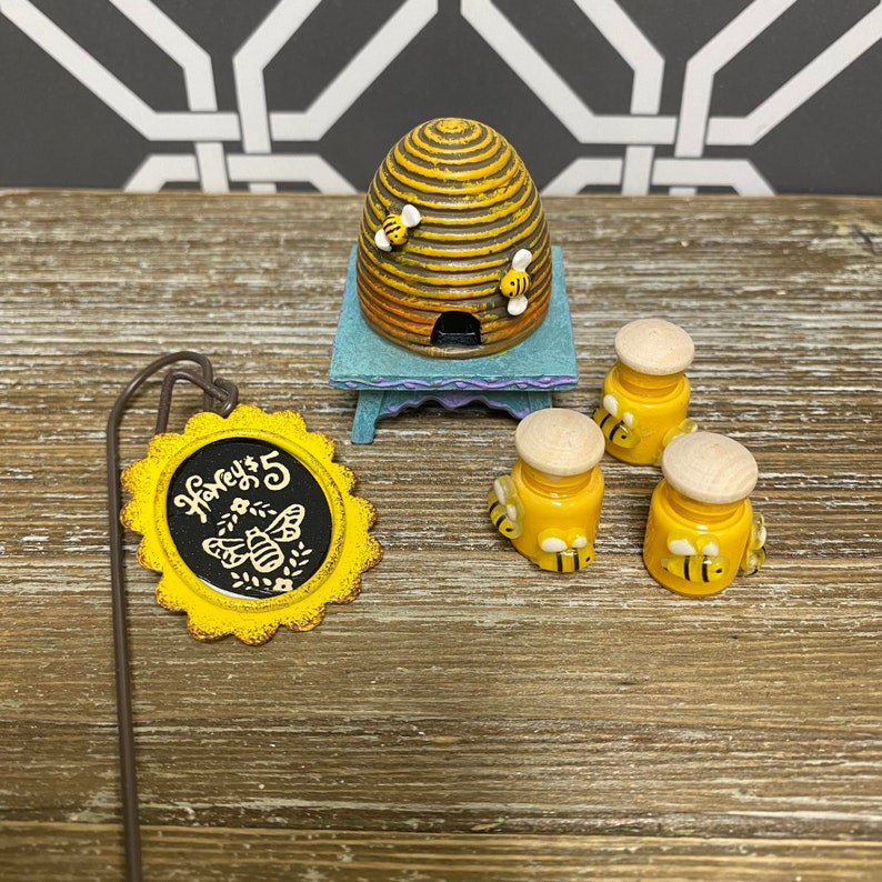 May include: A miniature beehive with three honey jars, each decorated with bees, and a sign that reads "Honey $5". The beehive is brown and yellow with a small entrance. The honey jars are yellow with white lids. The sign is yellow with black lettering and a bee illustration.