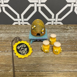 May include: A miniature beehive with a sign that reads "Honey's" and three small honey jars with bees on them. The beehive is brown and yellow with a blue base. The jars are yellow with white lids and have bees on them. The sign is yellow with black lettering.