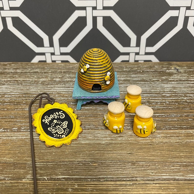May include: A miniature beehive with a sign that reads "Honey's" and three small honey jars with bees on them. The beehive is brown and yellow with a blue base. The jars are yellow with white lids and have bees on them. The sign is yellow with black lettering.