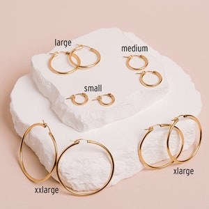 May include: A collection of gold hoop earrings in various sizes, including small, medium, large, xlarge, and xxlarge. The earrings are displayed on a white, textured surface against a soft pink background. The sizes are labeled with text.