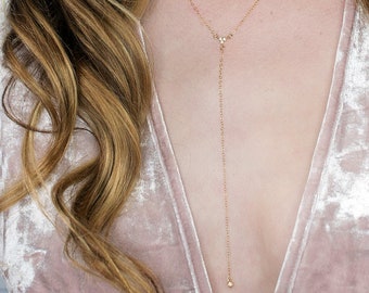 May include: A gold-toned layered necklace set featuring a short choker chain with small circular links and a longer Y-shaped lariat necklace with a delicate chain drop. The jewelry is worn against a light pink velvet top with a deep V-neckline.
