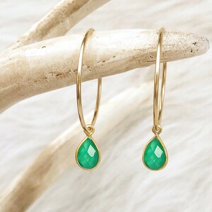 May include: A pair of gold hoop earrings with teardrop-shaped, faceted green gemstones. The earrings are displayed against a light-colored, textured background, showcasing their delicate design and vibrant color. The gemstones are approximately 0.5 inches in length.