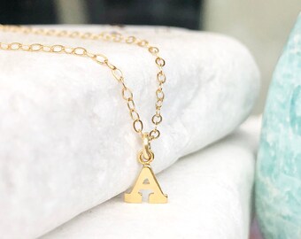 May include: A gold-coloured necklace featuring a small, capital letter A pendant suspended from a delicate, thin chain. The pendant has a smooth, polished finish and is attached to the chain by a small circular jump ring.