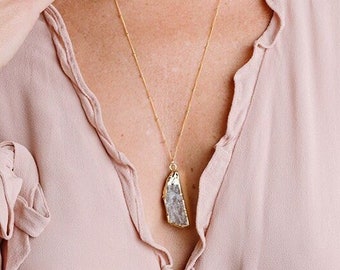 Peut inclure: Une femme portant un chemisier rose clair avec un collier en chaîne dorée et un grand pendentif en pierre grise et blanche irrégulière.