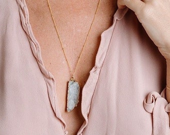 Peut inclure: Une femme porte un chemisier rose clair croisé et un collier en or avec un grand pendentif en pierre rectangulaire de couleur claire. Elle porte également un bracelet en or avec un motif de cœur. L'arrière-plan est de couleur neutre unie.