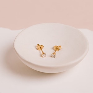 May include: A pair of gold stud earrings with small clear stones. The earrings are displayed on a white ceramic dish.