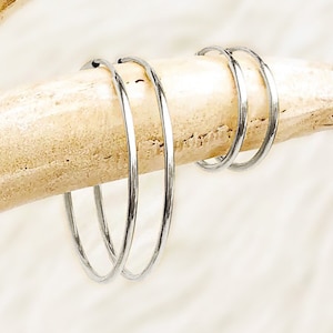 May include: A close-up shot of several silver hoop earrings. The earrings are thin and circular, and they are resting on a light-colored, textured surface. The background is a soft, white, blurred texture.