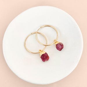 May include: A pair of gold hoop earrings with raw ruby gemstone drops. The earrings are displayed on a white, circular dish against a soft pink background. The ruby stones are a deep red color.