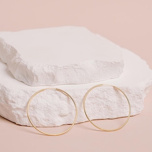 May include: Two gold-colored hoop earrings are displayed on a white, textured, stone-like surface against a soft pink background. The earrings are simple, circular, and have a thin profile. The overall aesthetic is minimalist and elegant.