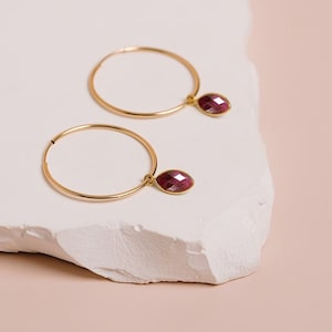 May include: A pair of gold hoop earrings with a dangling red gemstone. The earrings are on a white surface.
