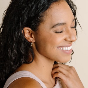 Peut inclure: Une femme aux cheveux longs et boucl&eacute;s fonc&eacute;s porte des boucles d'oreilles cr&eacute;oles en or. Les boucles d'oreilles sont de taille moyenne et ont une fermeture &agrave; levier. Le mod&egrave;le sourit et regarde vers le bas. Le texte "model is wearing medium leverbacks" est visible en bas de l'image.
