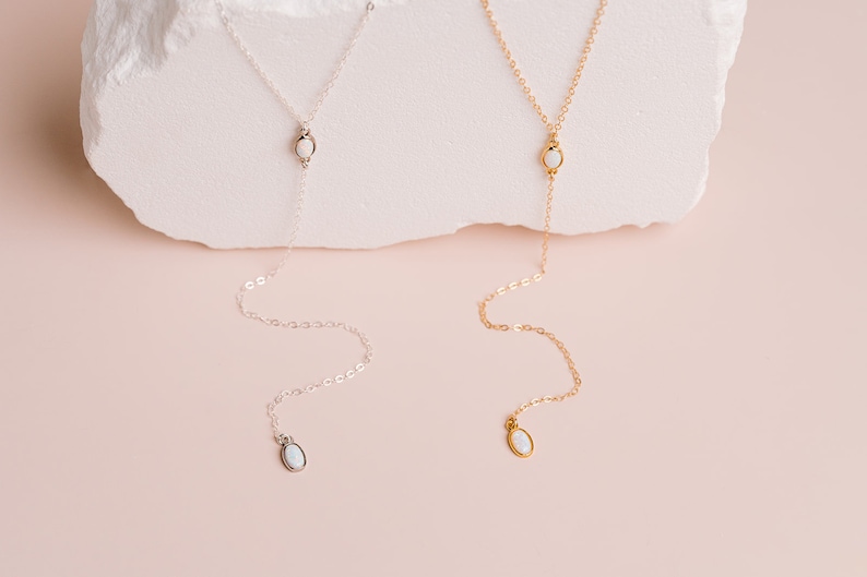 Puede incluir: Dos collares tipo lazo, uno en tono plateado y otro en tono dorado, cada uno con una cadena delicada, un peque&ntilde;o acento de piedra blanca ovalada en el centro y un colgante de piedra blanca ovalada a juego al final de la cadena.