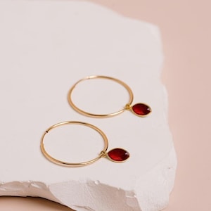 May include: A pair of gold hoop earrings, each featuring a single dangling marquise-cut red gemstone charm. The thin, circular gold hoops are polished and minimalist, with the deep red, faceted stones suspended from small gold loops.