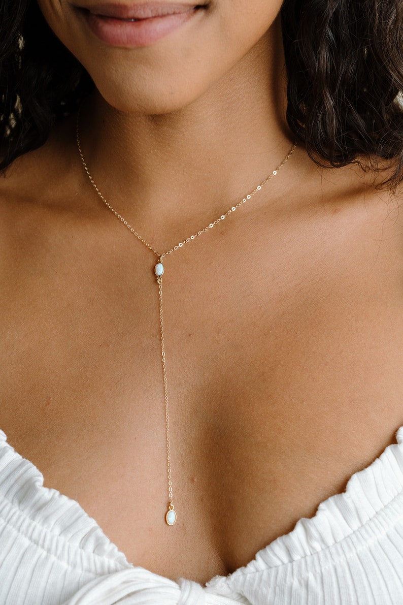 Puede incluir: Un collar tipo lariat en tono dorado con una cadena fina y dos peque&ntilde;as piedras blancas ovaladas de aspecto opalino. Una piedra se sit&uacute;a en la uni&oacute;n central, mientras que la segunda cuelga al final de la cadena vertical que cae.