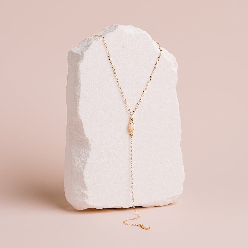Puede incluir: Un collar tipo lariat en tono dorado con una cadena de eslabones finos y dos peque&ntilde;as piedras ovaladas de color blanco similar al &oacute;palo. Una piedra se sit&uacute;a en el punto central de ca&iacute;da y la otra sirve como remate al final de la cadena.